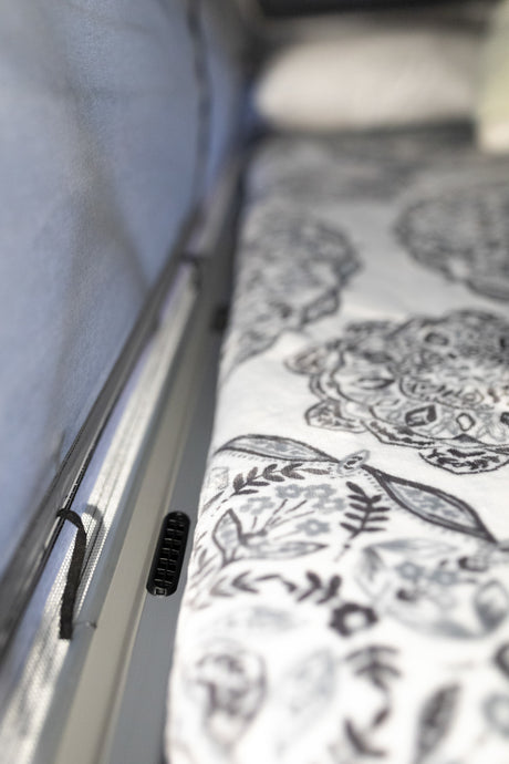 Inside an RV rental: A patterned bed with white linens borders a light wall, featuring a vent and a mesh screen.