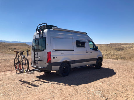 Silver RV rental van with mountain bikes on a rack, parked in a vast, sunny desert landscape.