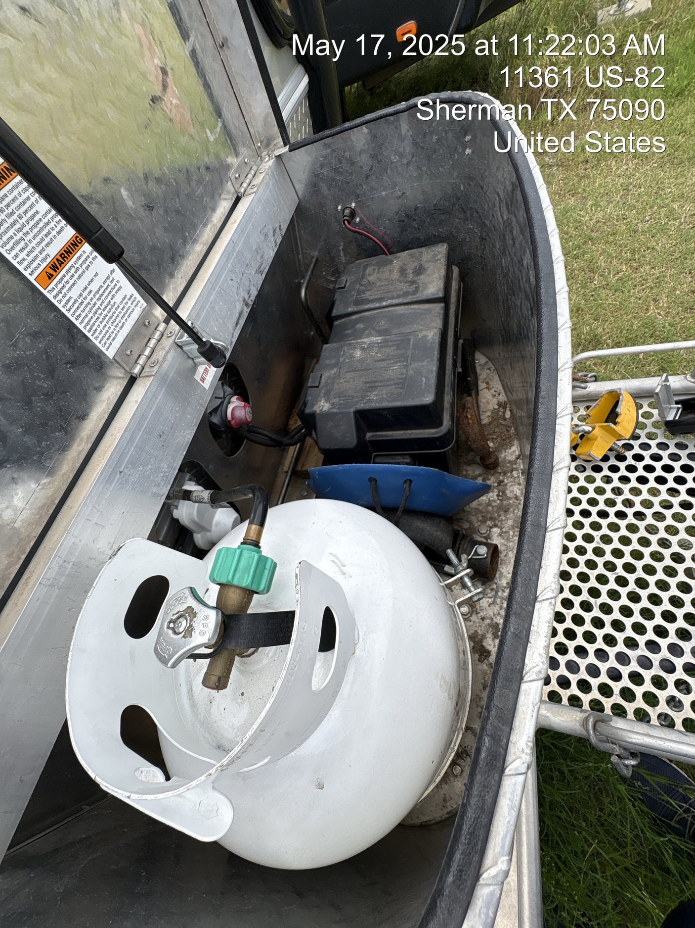 An RV rental's open storage shows a white propane tank, black battery box, and safety warnings for essential use.