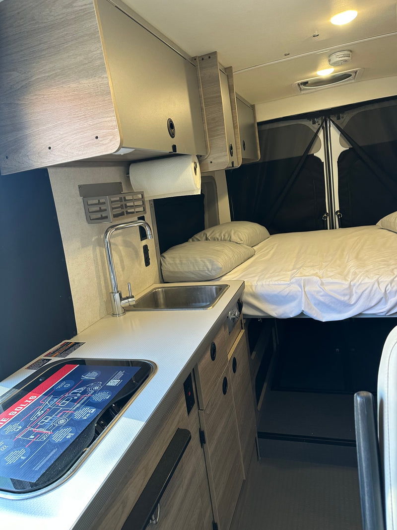 Bright RV rental interior featuring a practical kitchen with sink/stove and a comfy bed. Essential amenities for travel. — photo 11