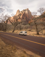 White camper van on a dark road in a valley with towering red mountains and bare trees, ideal for an RV rental trip.