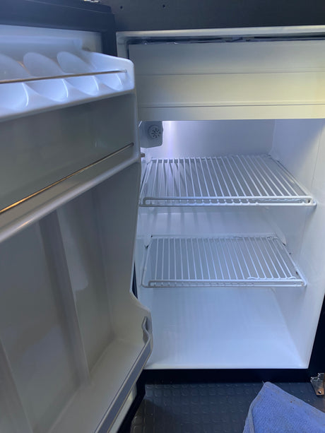 Empty, clean mini-fridge in an rv rental, with door open showing wire shelves and interior light. Ready for your trip!