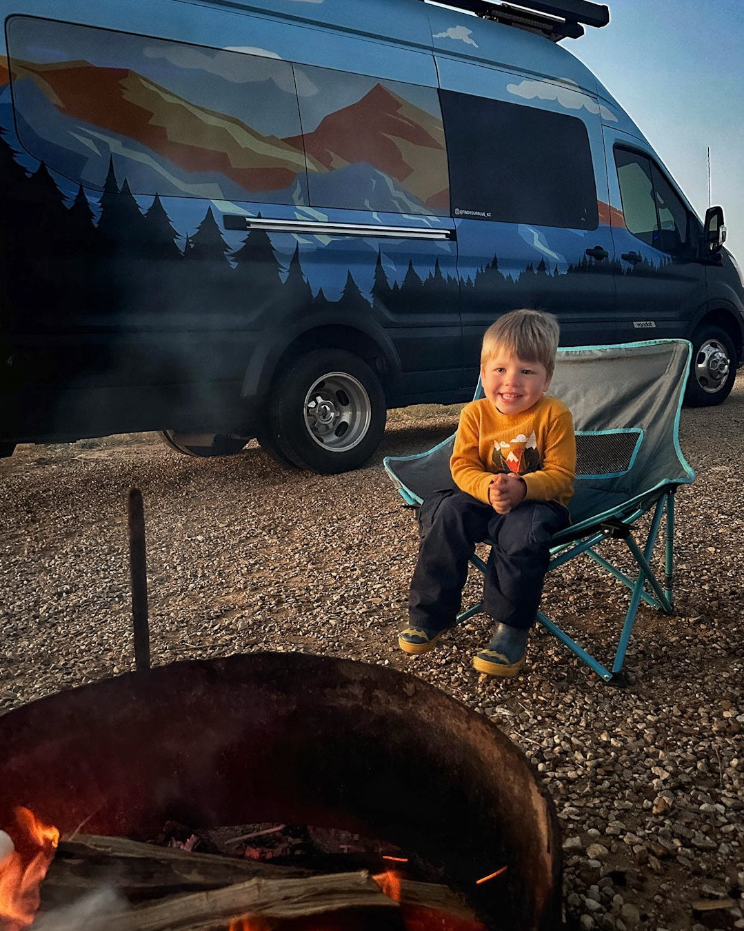 Camping fun! A happy child sits by a campfire with a custom RV rental van in the background, painted with mountains and trees.