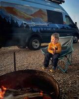Camping fun! A happy child sits by a campfire with a custom RV rental van in the background, painted with mountains and trees.