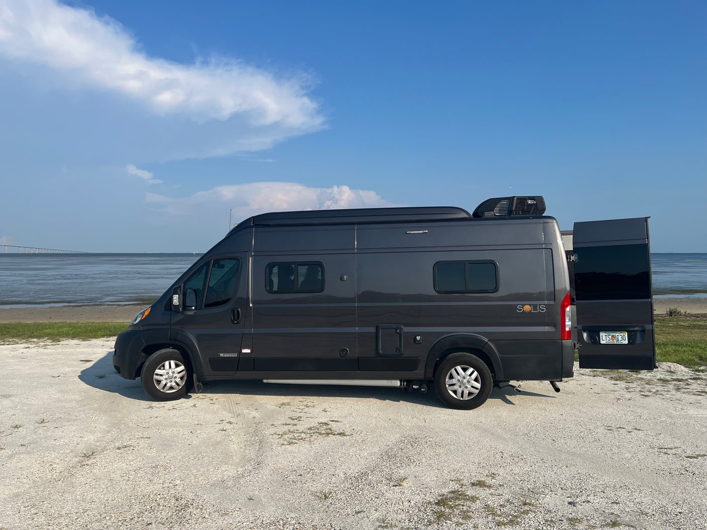 RV rental: Winnebago Solis camper van by the water with a long bridge, under a clear blue sky.
