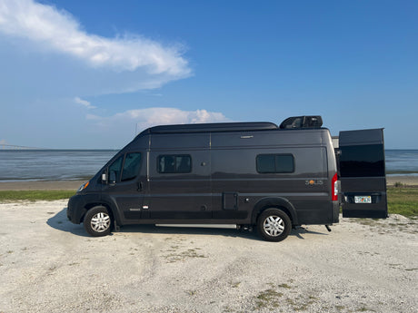 RV rental: Winnebago Solis camper van by the water with a long bridge, under a clear blue sky.