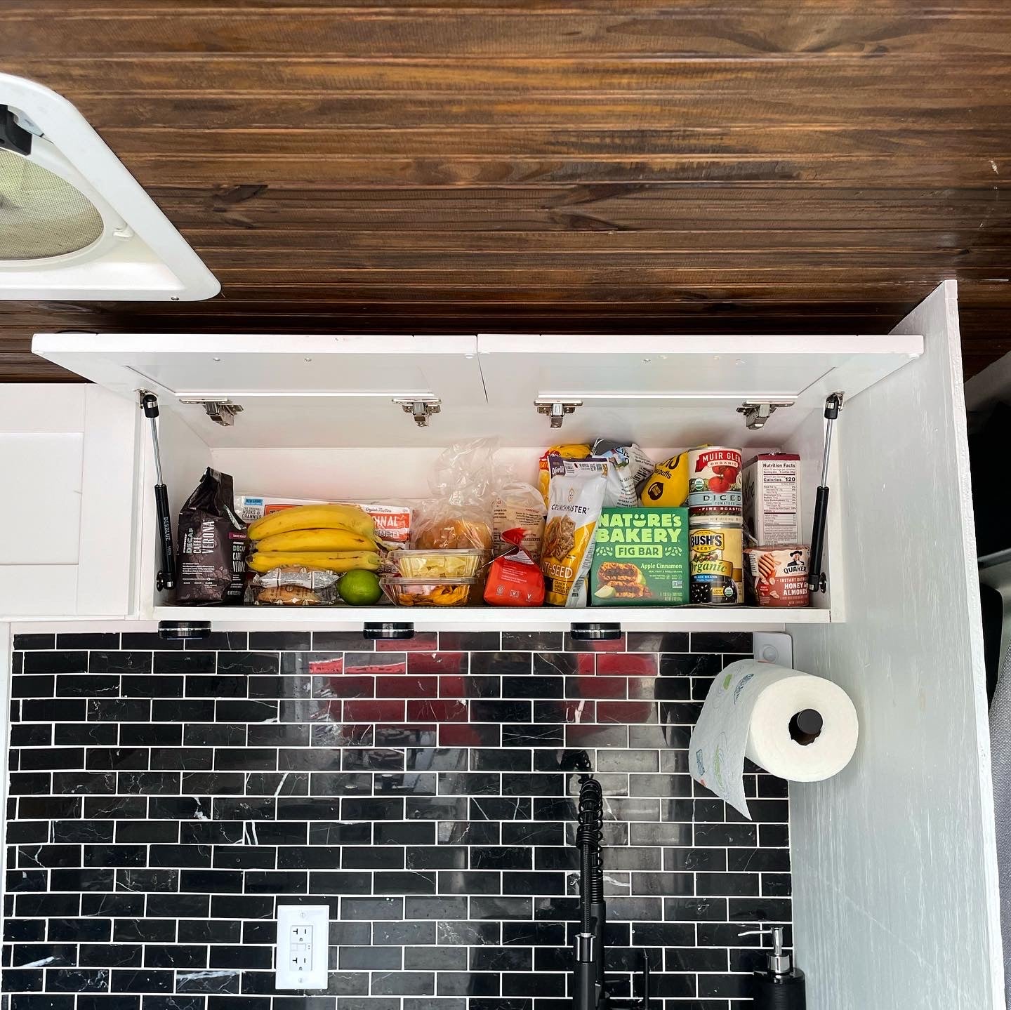 Compact RV rental kitchen features an open cabinet filled with food, black subway tile backsplash, and a paper towel holder.