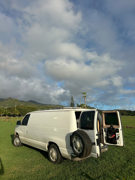 White camper van, ideal for an RV rental, parked in a grassy field with open rear doors, tropical mountains, and cloudy skies.