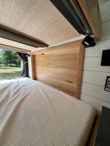 Cozy RV rental interior: Light wood headboard, quilted bed, white walls, and a window view of lush nature.