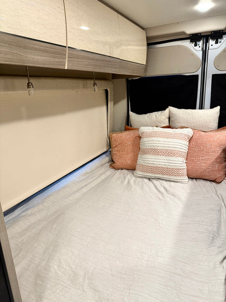 Cozy RV rental sleeping area: a comfortable bed with light bedding, accent pillows, overhead cabinets, and lights.