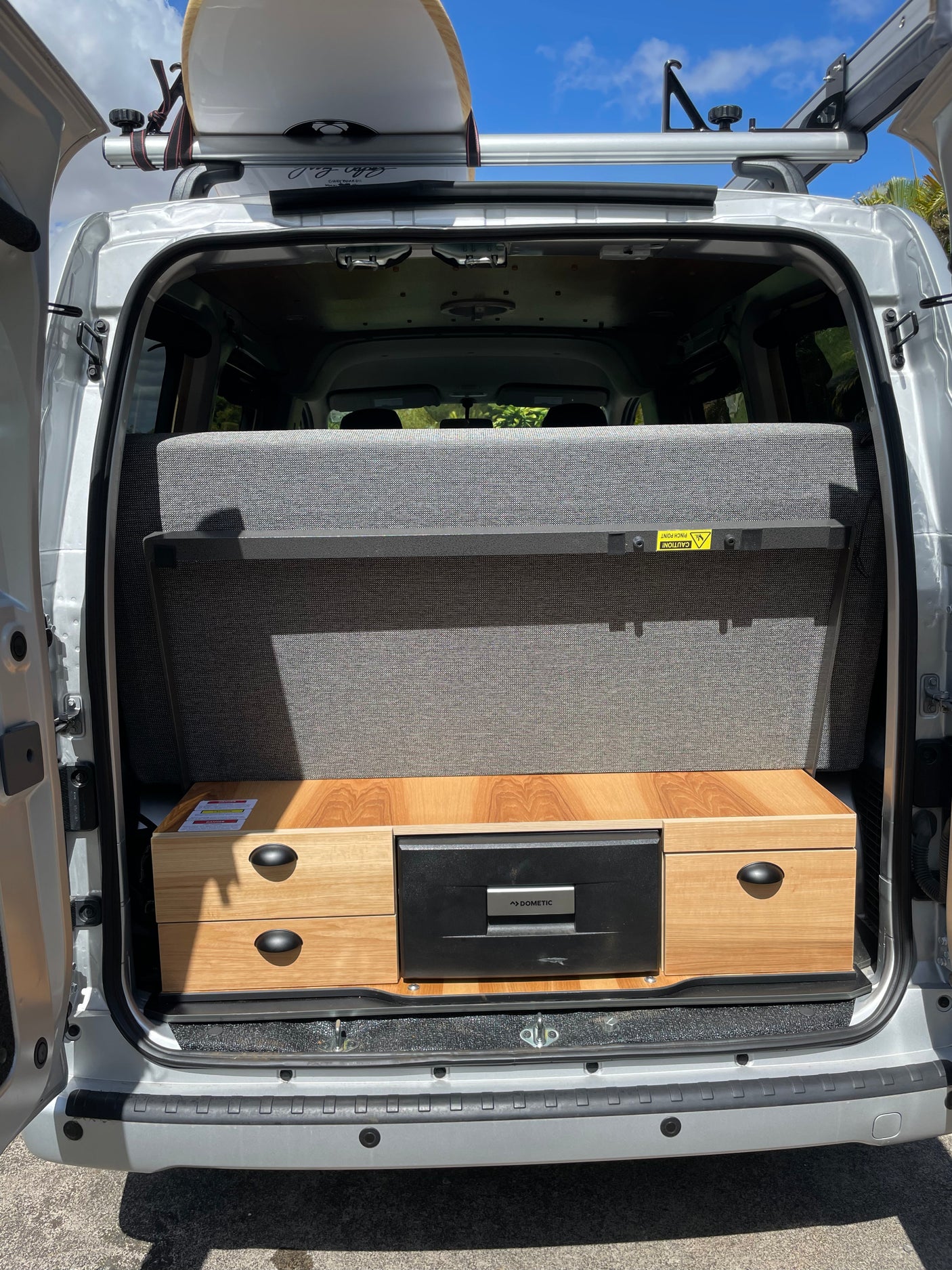 Rear view of an RV rental: Silver campervan interior with gray pull-out bed, wooden storage, Dometic fridge, and surfboard on roof rack.