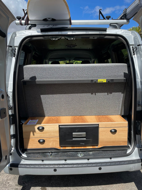 Rear view of an RV rental: Silver campervan interior with gray pull-out bed, wooden storage, Dometic fridge, and surfboard on roof rack.