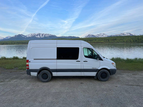 White RV rental van by a scenic lake with snow-capped mountains. Ideal for vanlife adventures & road trips.