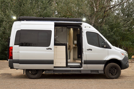 Side view of a silver CanaK camper van, great for rv rental trips. Its door is open, revealing the interior & roof awning lights.