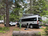A Tellaro RV rental and white Tesla Model Y charging at a treed campsite. A man stands between the vehicles.