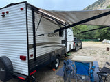 An RV rental, a white travel trailer with an extended awning, is set up at a dirt campsite with a pickup truck and chairs.