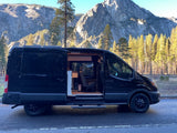 Black RV rental van with open side door showing converted interior, parked against a backdrop of majestic mountains and tall pines.
