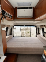 RV rental interior featuring a comfy bed, LG AC unit, and a scenic beach view from the rear of the camper van.