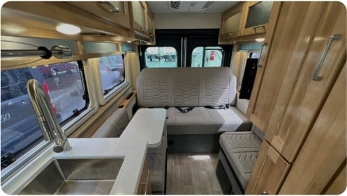  and large windows revealing a rainy day.|Spacious RV rental dinette: comfortable seating with textured cushions & table. Ideal camper interior.|Inside this RV rental: a modern kitchen with sink — photo 7