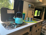 RV rental interior: Compact kitchen with stove, sink, kettle, and mugs. Features window view of trees and sleeping area.