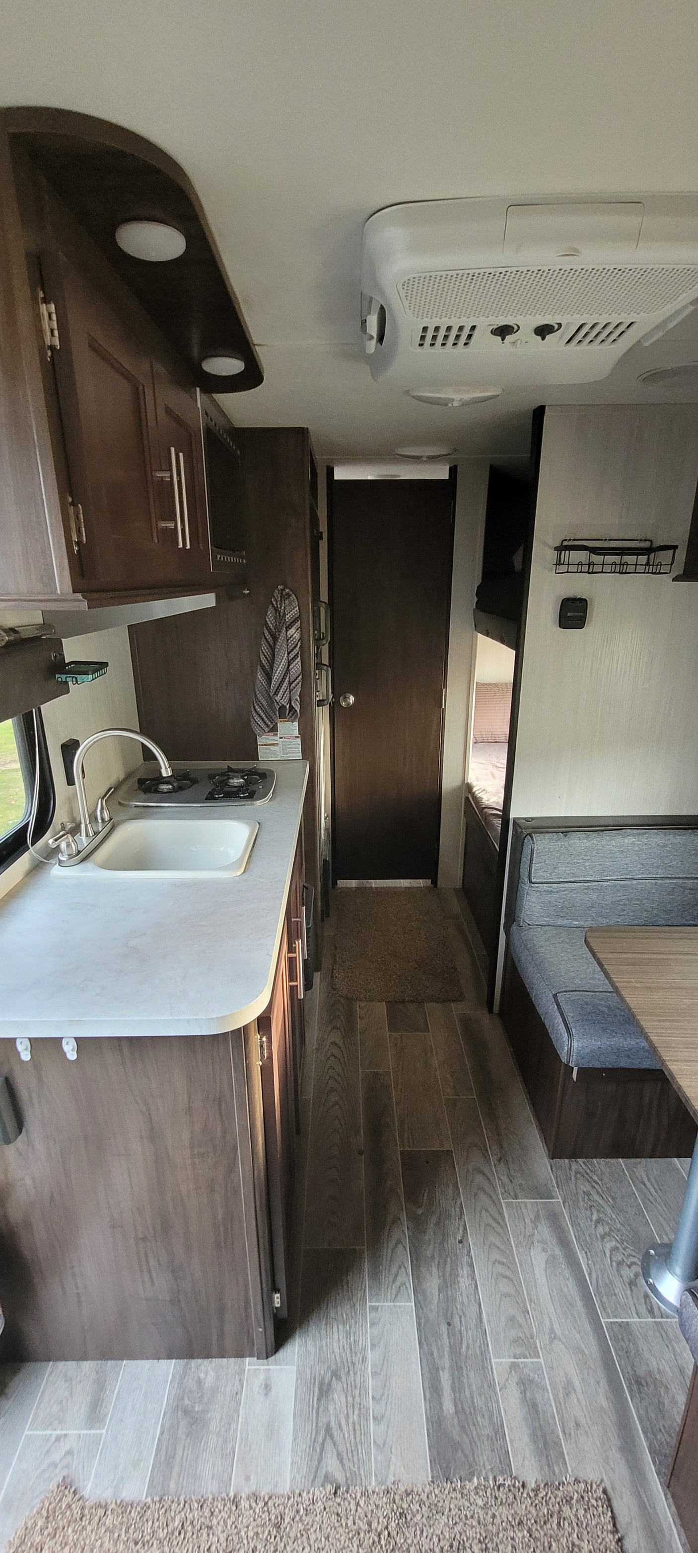 RV rental interior: compact kitchen with sink & stove, dark wood cabinets, wall AC unit, and wood-look floor.