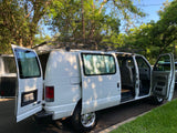 White Ford Econoline RV rental with open doors & roof rack, parked under trees. Ready for adventure!