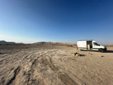 A white RV rental camper van parked in a wide-open, dry desert area with distant mesas. A rock fire pit is nearby.