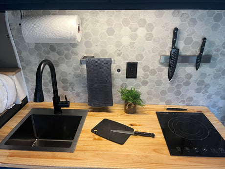 Compact RV rental kitchen: sleek black sink/faucet, induction cooktop, hex tile backsplash, paper towels, and knives.