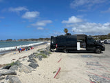 Black rv rental camper van on a sandy beach. People relax by ocean waves under a blue sky. Embrace van life adventure!