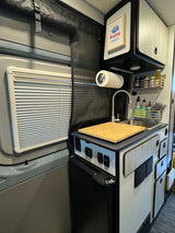 Compact RV rental interior showing a small kitchen with sink, fridge, overhead cabinets, and window blinds.