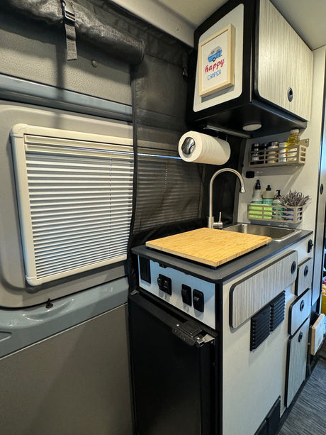 Compact RV rental interior showing a small kitchen with sink, fridge, overhead cabinets, and window blinds.