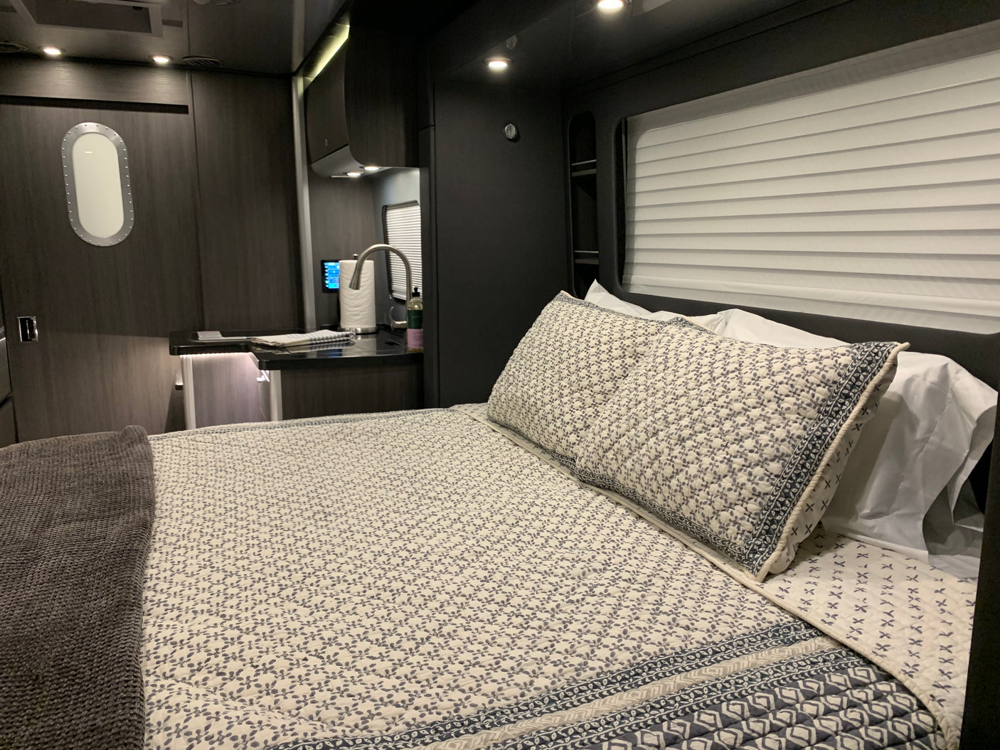 Modern rv rental interior: bedroom with patterned bedding & grey blanket, compact kitchen area, and unique oval window.