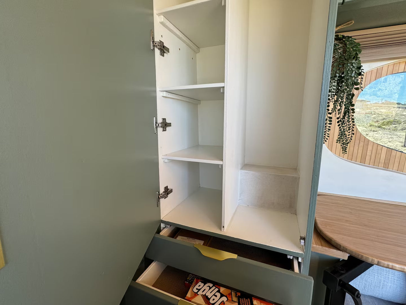 Open sage green cabinet in an RV rental reveals white shelves & drawers. Efficient storage for compact living by window.