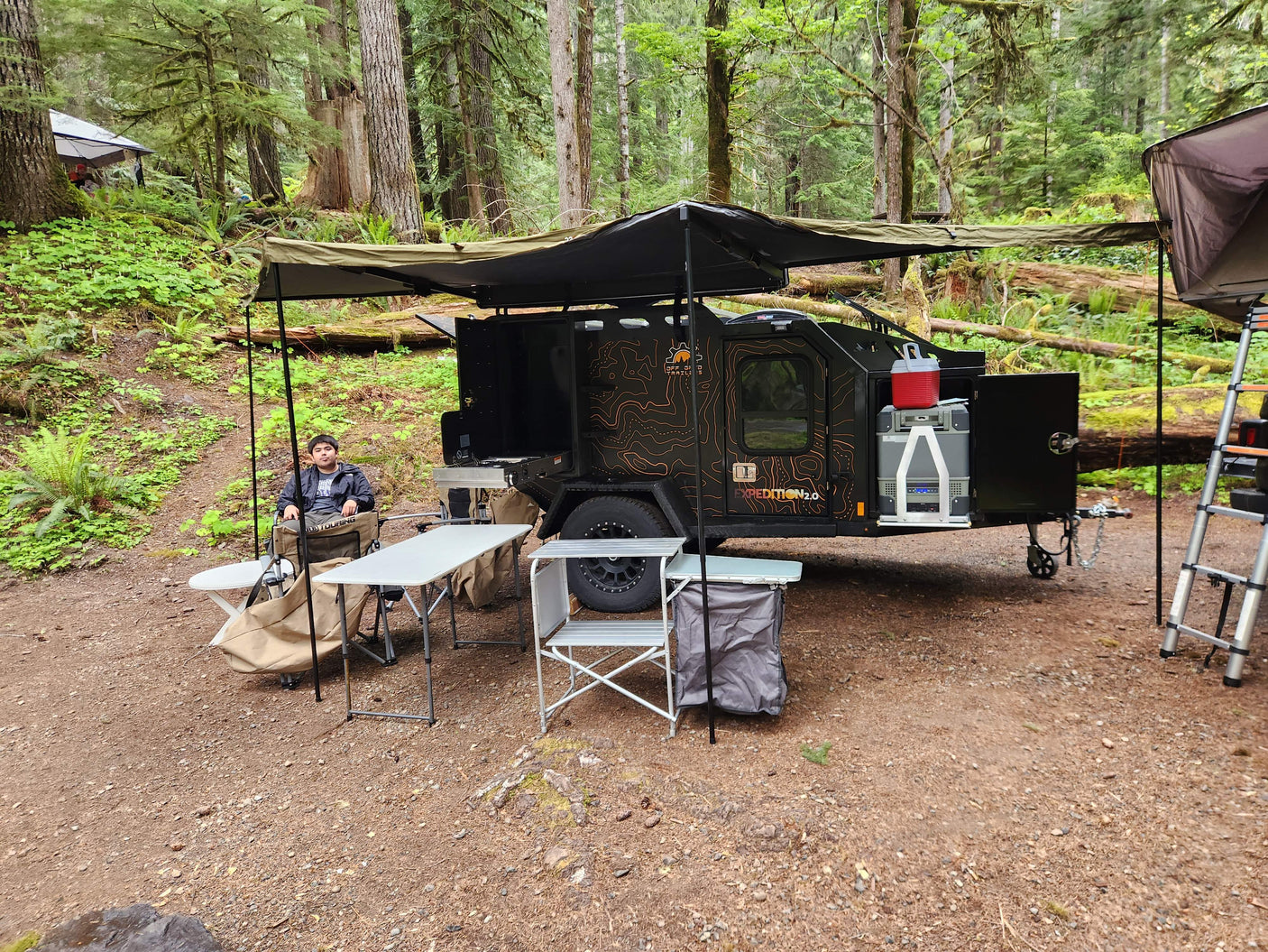 A spacious Off Grid Trailers Expedition 2.0 rv rental set up for comfort in a lush forest camp, with a person relaxing.