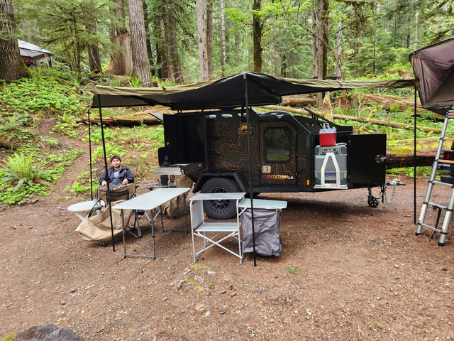 A spacious Off Grid Trailers Expedition 2.0 rv rental set up for comfort in a lush forest camp, with a person relaxing.