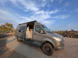 Grey Mercedes Sprinter RV rental parked on a paved lot with its side door open, revealing a functional kitchenette and interior.