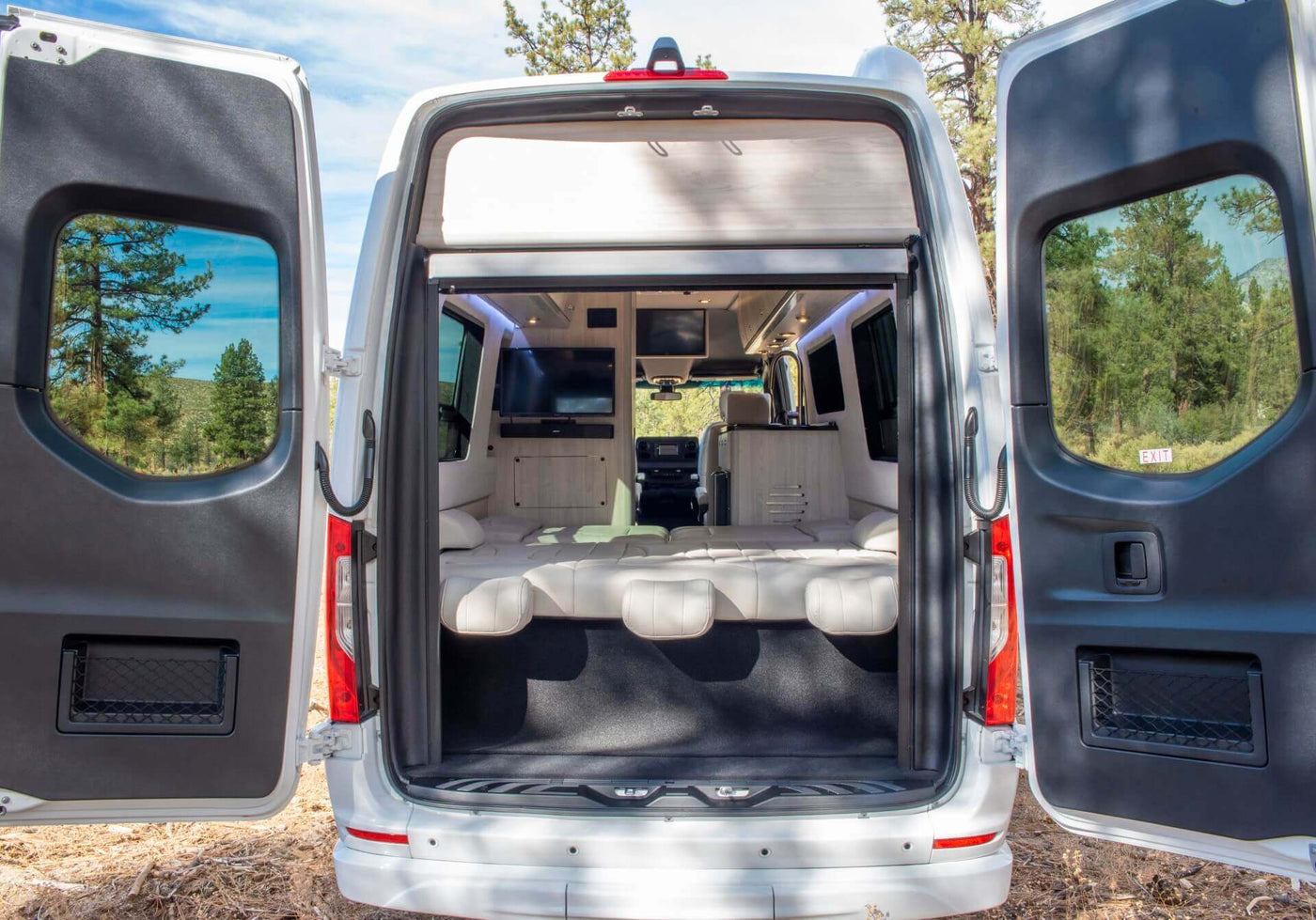  and a stunning lake view window.|A modern white Mercedes-Benz Sprinter RV rental parked on a dirt path in a peaceful pine forest setting.|Enjoy a luxury RV rental! View the spacious Mercedes-Benz Sprinter interior with an overhead TV and scenic window views.|Spacious RV rental interior: luxurious seating — photo 18