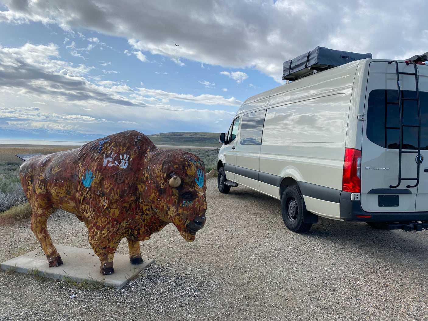 An RV rental Sprinter van parked by a colorful bison statue, overlooking a lake and mountains. Great outdoor travel.
