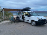 White RV rental camper van with awning & chairs by a lake. Enjoying an evening camp setup.