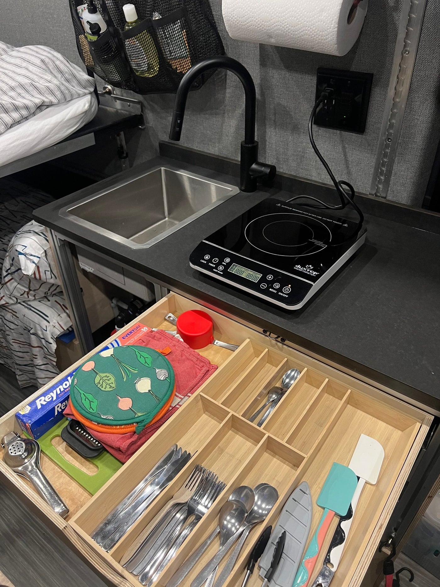 A compact RV rental kitchen features a sink, an induction cooktop, and an open drawer with organized cooking utensils and foil. — photo 17