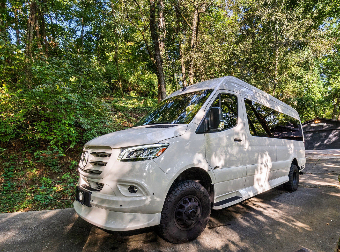 Explore nature with this white Mercedes Sprinter adventure van, ideal for an RV rental. Parked on a sunny day amongst lush trees.
