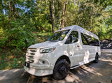 Explore nature with this white Mercedes Sprinter adventure van, ideal for an RV rental. Parked on a sunny day amongst lush trees.