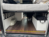 Inside a spacious RV rental camper van: elevated bed, wood floor, and integrated storage, with measuring tape on floor.