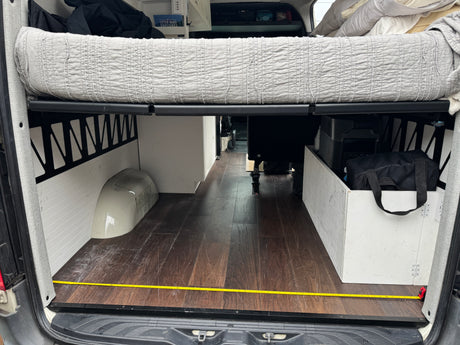 Inside a spacious RV rental camper van: elevated bed, wood floor, and integrated storage, with measuring tape on floor.