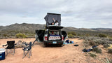 Desert camping with an RV rental: An SUV with a pop-up rooftop tent sits amidst mountains. People relax, enjoying the outdoor adventure.