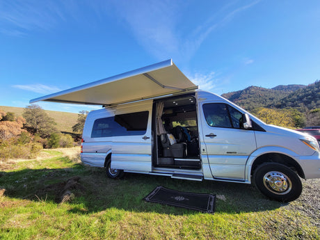 An RV rental set up in a vibrant natural landscape. The silver camper van's awning is out, and door is open on green grass.