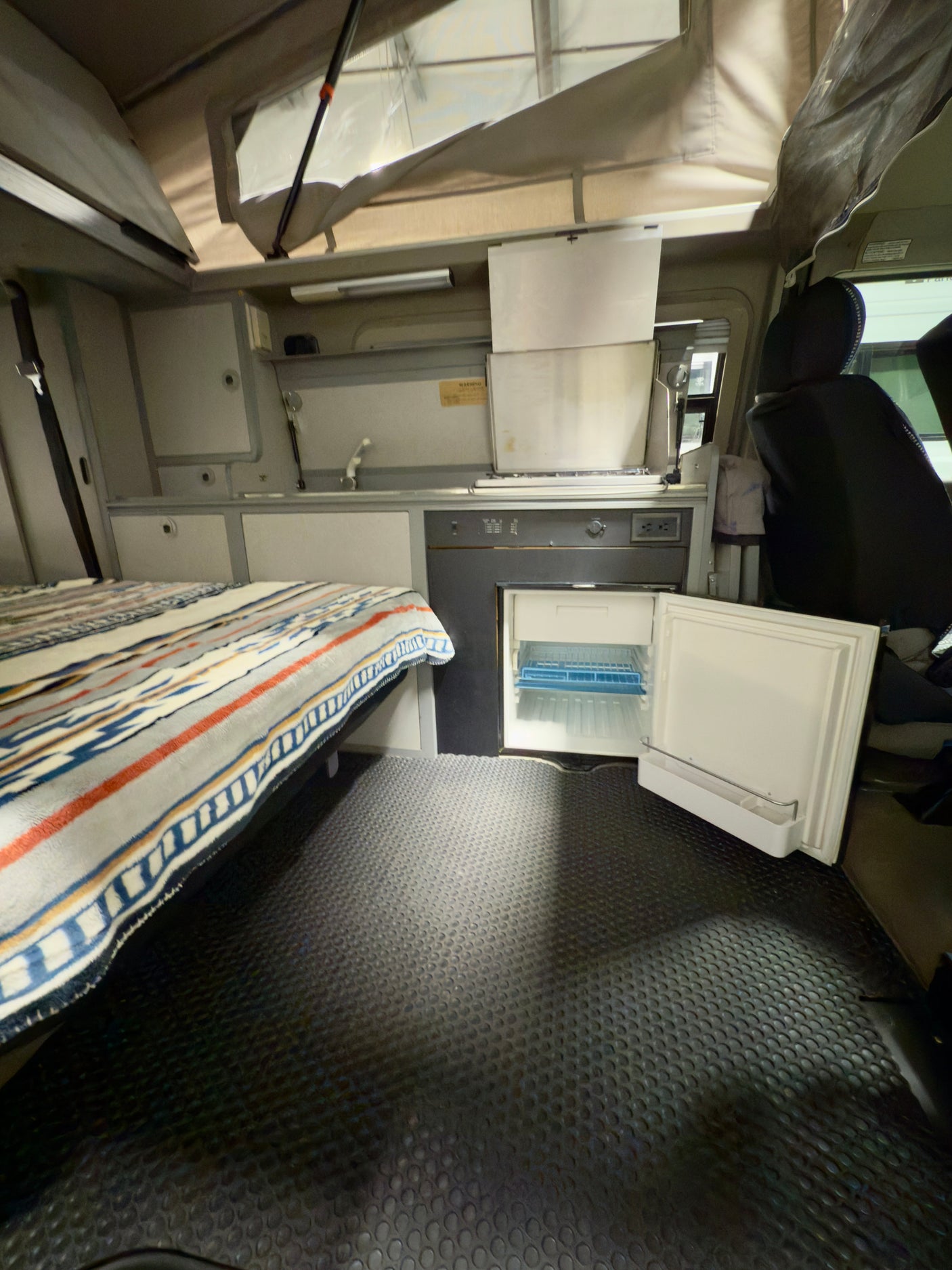 RV rental interior shows a cozy bed, kitchenette with open fridge, and pop-top roof offering ample space.