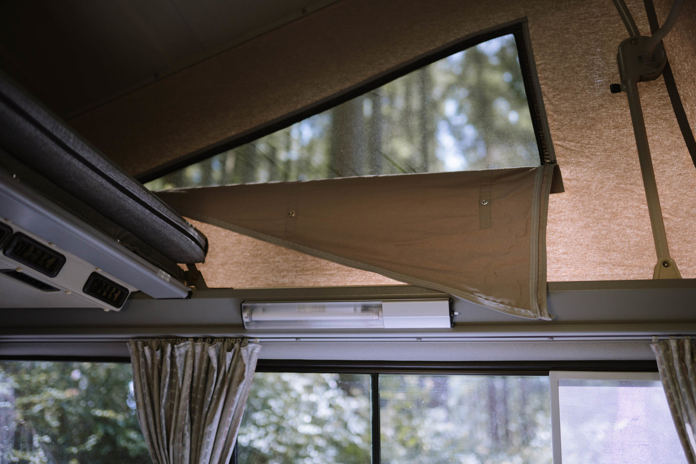Inside an RV rental camper: a raised pop-top tent with a mesh window views of trees, and a lower window with curtains.
