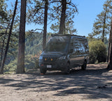 A dark Mercedes Sprinter camper van, ideal for an RV rental, is parked among tall pine trees in a sunny forest clearing.
