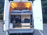 White RV rental van's open rear reveals a custom wood interior with bed, bike storage, helmet, & gear. Warm lights glow.